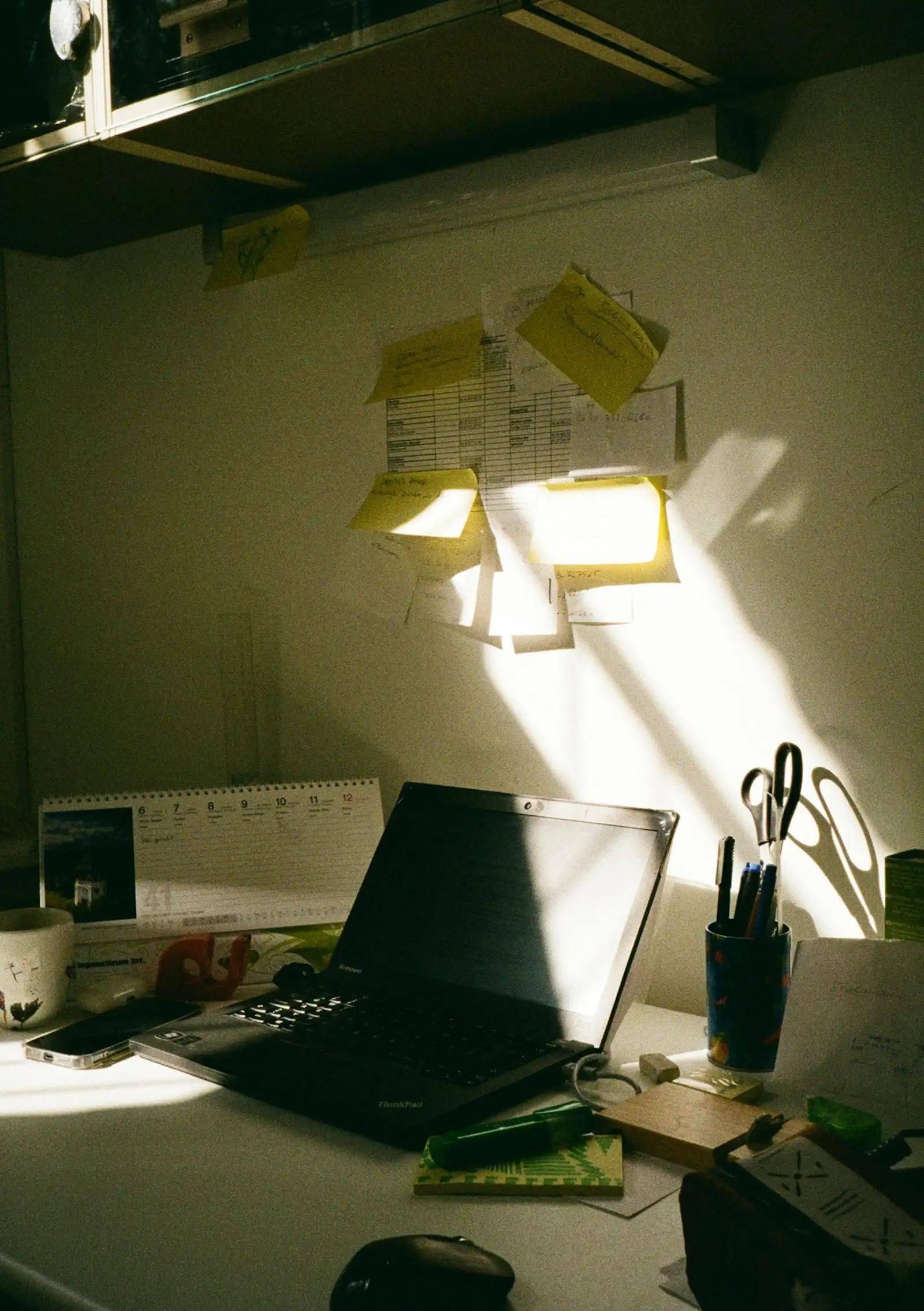 Arbeitsplatz mit Laptop, Notizzetteln an der Wand und Stiftebecher im Sonnenlicht